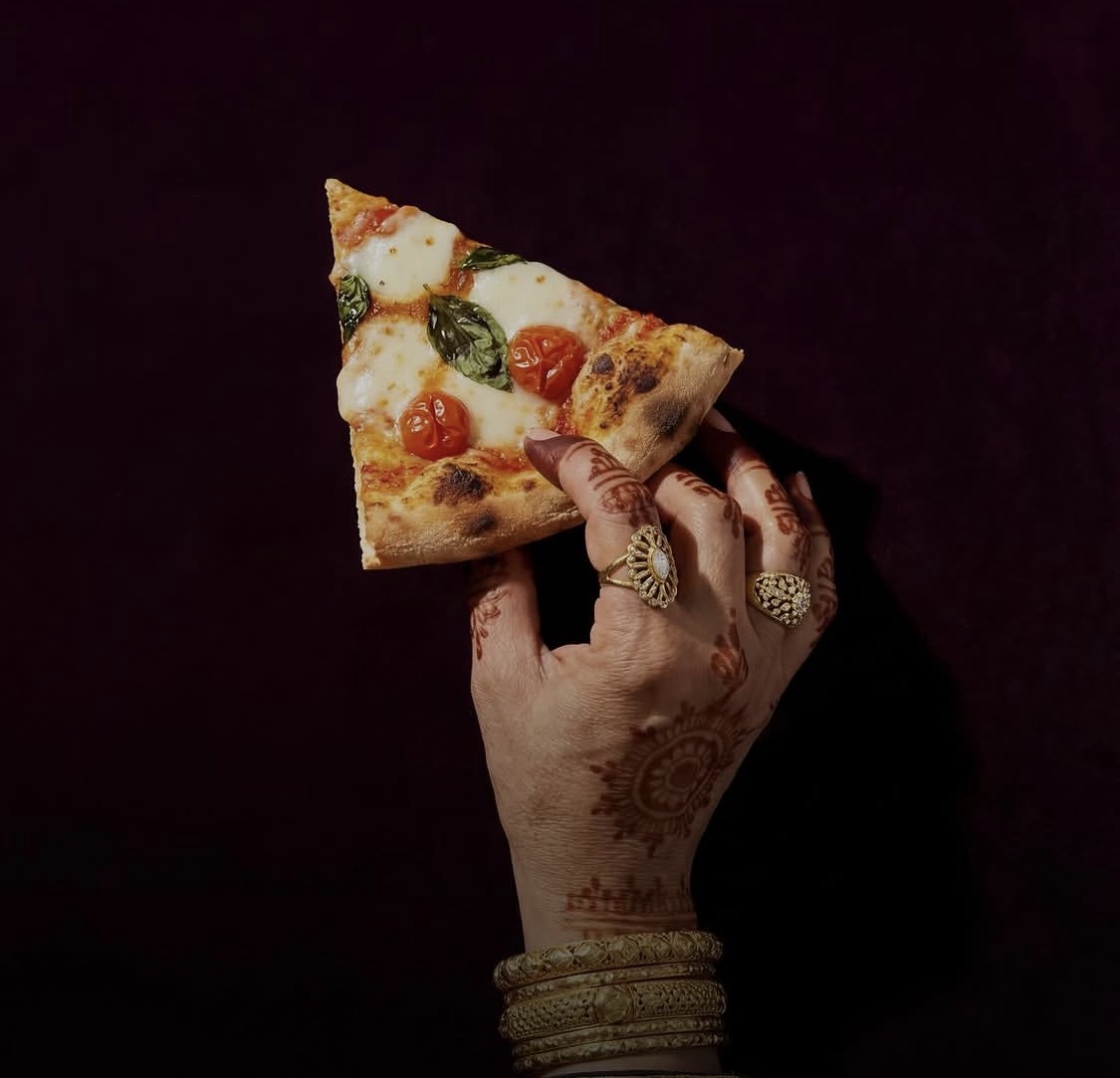A hand adorned with henna and gold rings holding a slice of margherita pizza